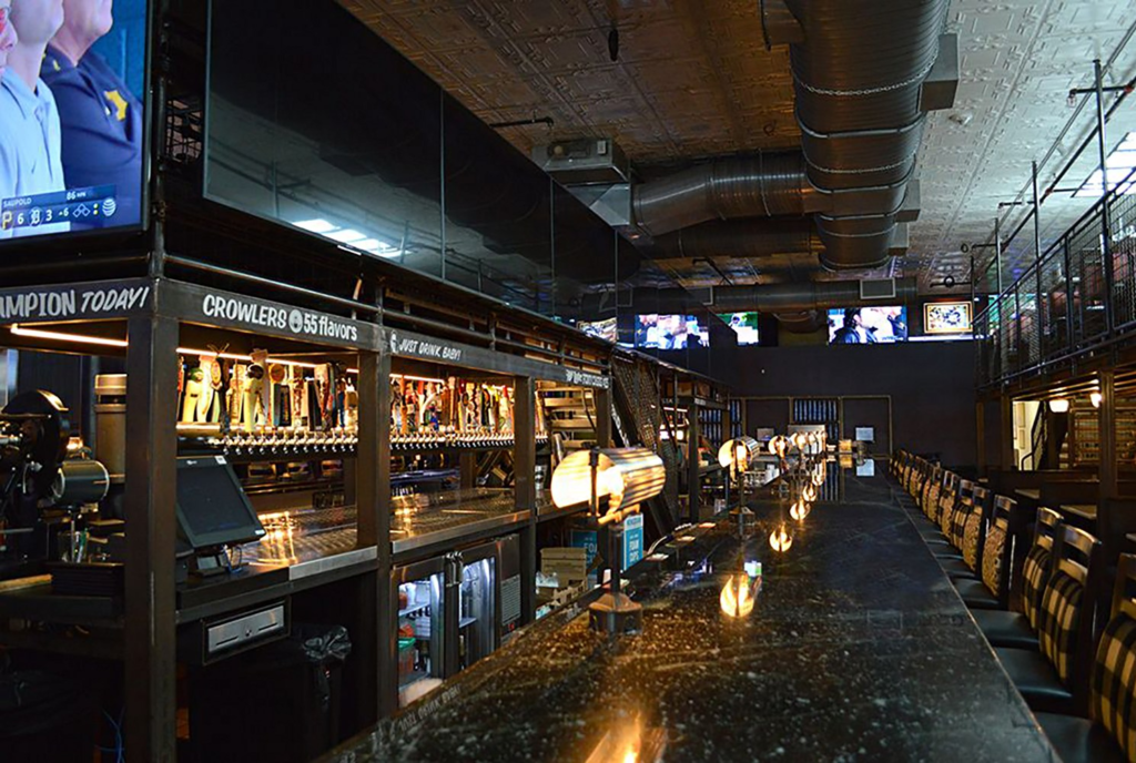 A dark bar with a long counter, library style lamps, rows of bottles on metal shelving, and TVs playing sports above.