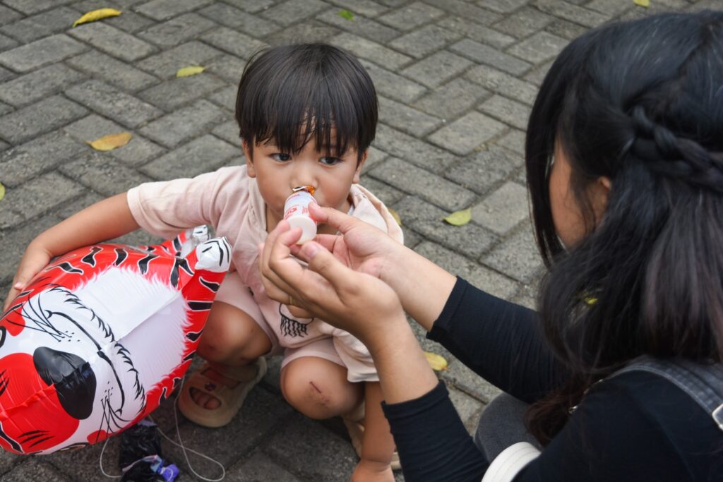 A woman helps a young boy drink a bottle of Yakult.