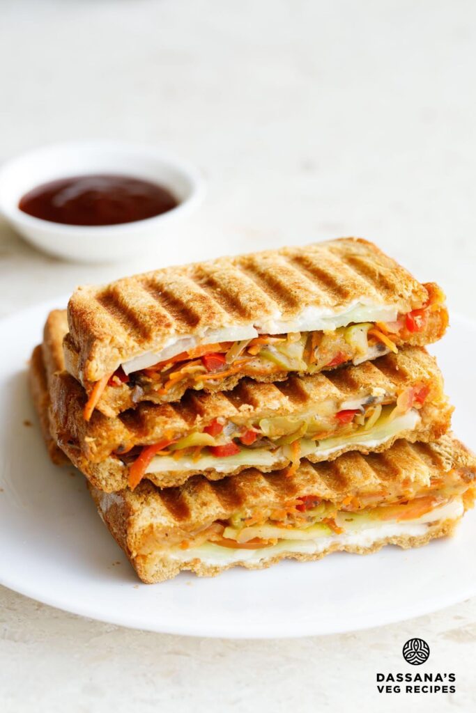 veg sandwich stacked on top with ketchup in a small bowl kept behind.