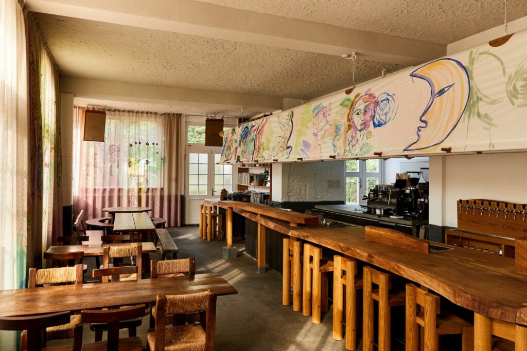 A restaurant interior with a long, natural wood bar, colorful mural above the bar, and curtains letting in light on wooden tables.