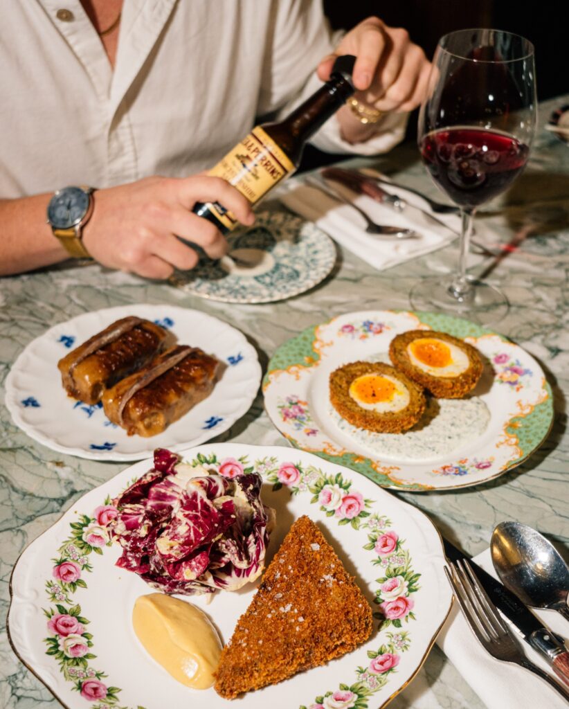 a spread of dishes at lord’s including crispy pig’s head served with radicchio, a scotch egg, and welsh rarebit. in the background, a diner grips a bottle of worcestershire sauce.