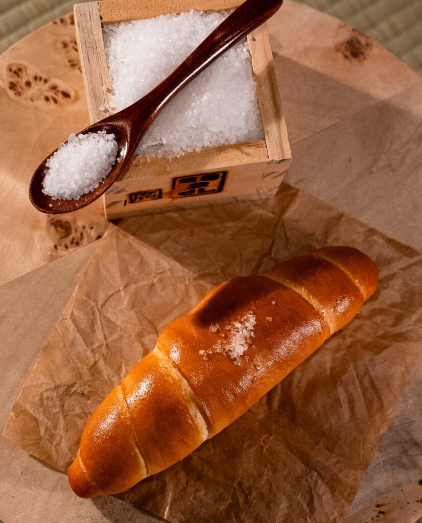 a roll of salt bread from Justin’s Salt Bread in NYC sits on a piece of brown parchment paper next to a wooden box of salt