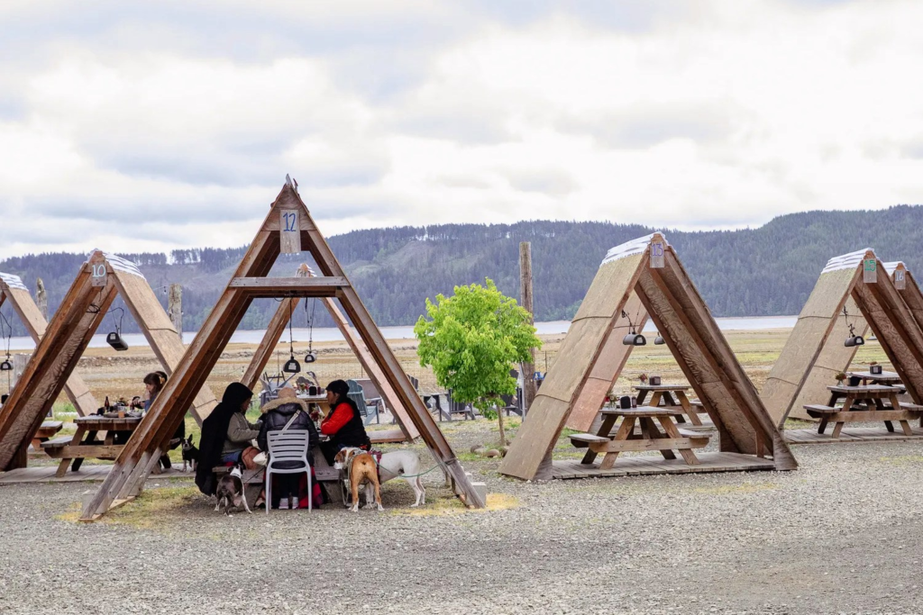 Tables set beneath A-frame wooden structures, set against a landscape of mountains and water.