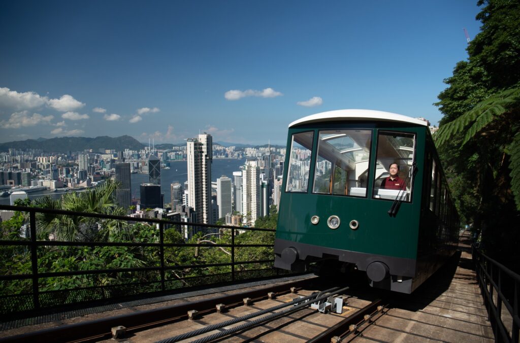 A tram climbs a hill with the skyscrapers of Hong Kong visible beyond.
