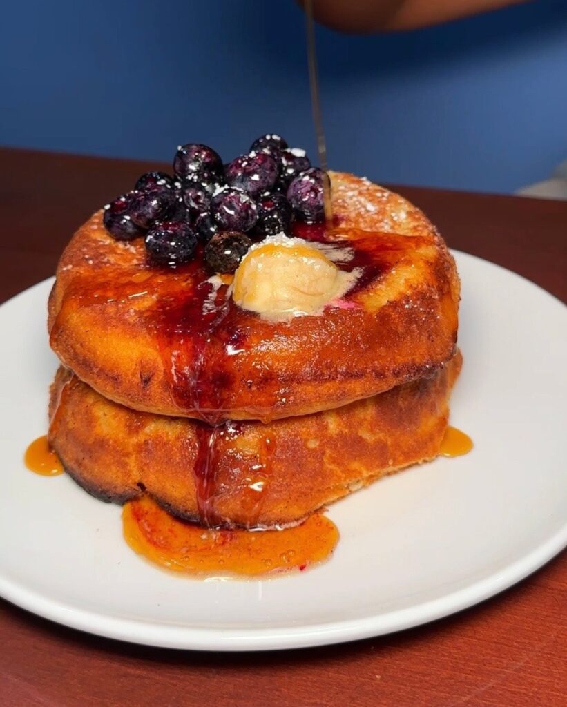 two thick pancakes on a white plate. the pancakes are topped with blueberries, butter, and syrup.