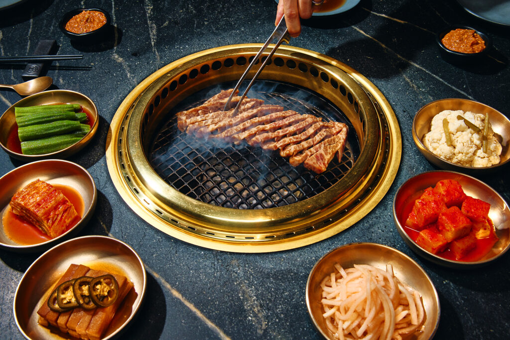 A server grills meat on a Korean in-table grill with banchan places all around.