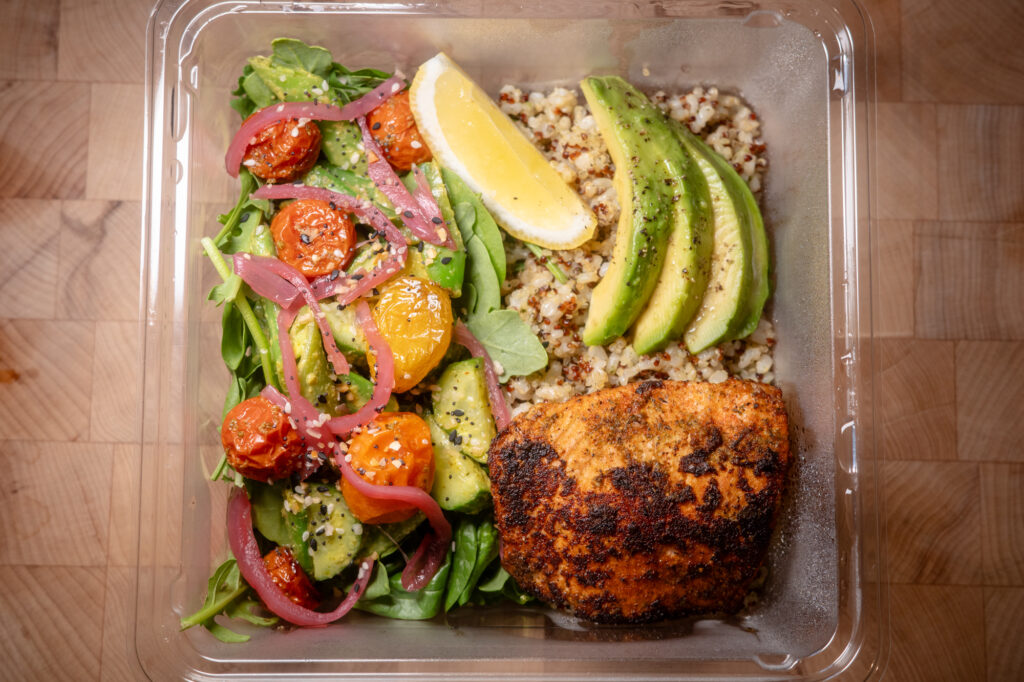 A takeout container featuring quinoa, salmon, avocado, and blistered tomato salad.
